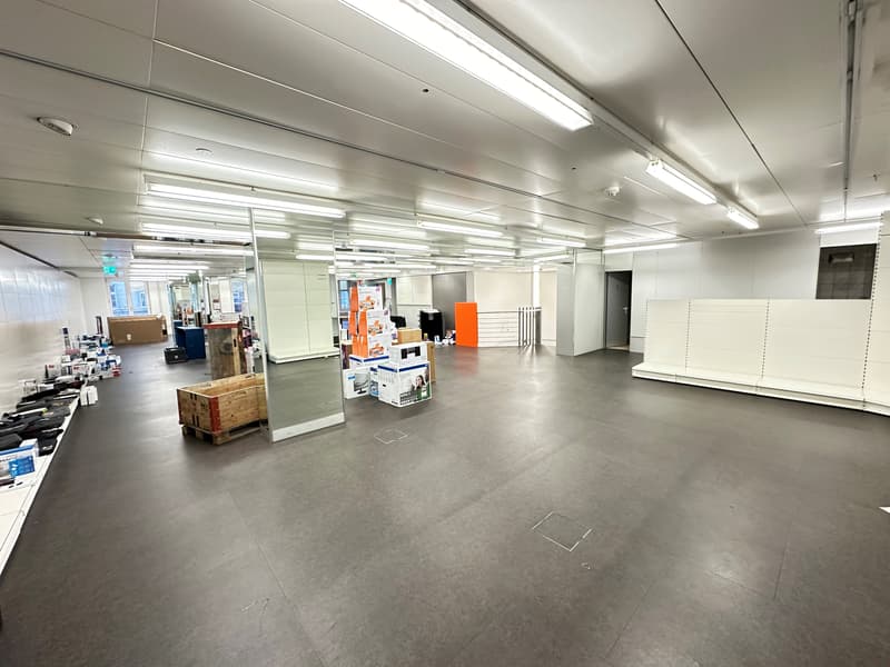 A wide open space with multiple fluorescent lights installed on the ceiling. There are shelves with some storage boxes on the left side of the room. Some storage boxes are also placed on the floor.