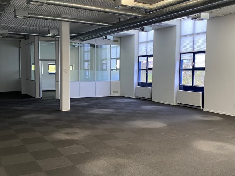 An open office space with a checkered floor, glass walls, windows with blinds, and pipes and lights on the ceiling.