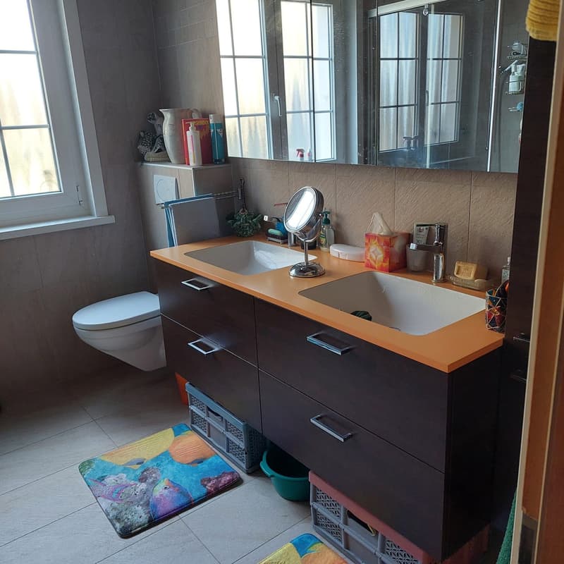 a bathroom with white tiles, a sink with two basins, a large mirror with storage boxes and a toilet.