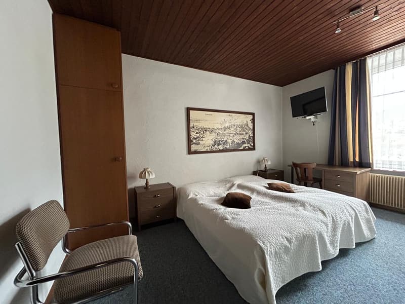 Spacious bedroom with wooden ceiling, white walls, large bed, nightstands, dresser, and a framed artwork on the wall. There is also a TV mounted on the wall and a chair in the corner.