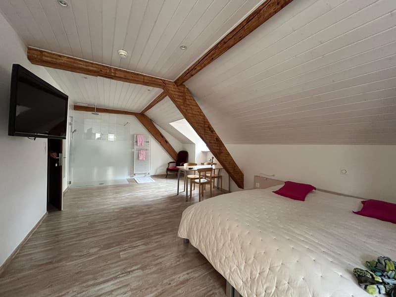 Spacious attic bedroom with wooden beams, white walls, hardwood flooring, large bed, and a seating area with a table and chairs near the window.