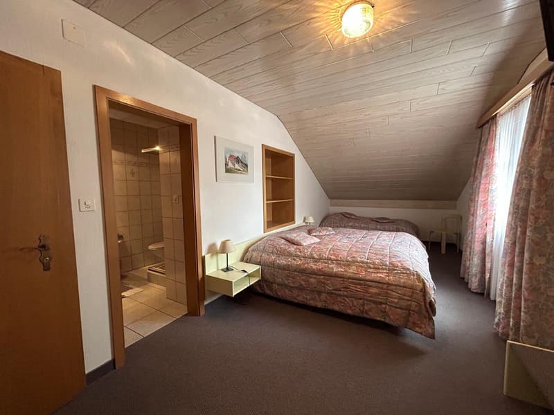 Attic bedroom with slanted wooden ceiling, large bed, bedside tables, en-suite bathroom, and curtained windows.