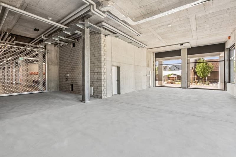 Large open commercial space with concrete floors, exposed ceiling, and brick walls. There is a metal security gate and a view of the exterior through the large windows.