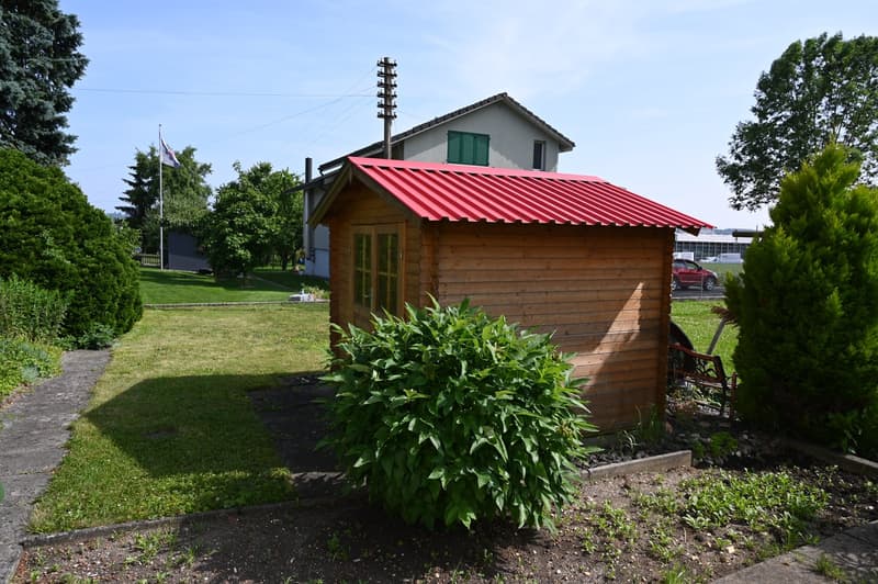 Small wooden house with red roof, door and window, flag, bench, trees, garden, and grass field