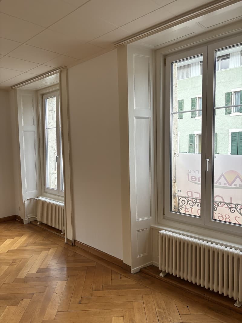 Wooden parquet floor, white walls, two windows, two radiators, open door