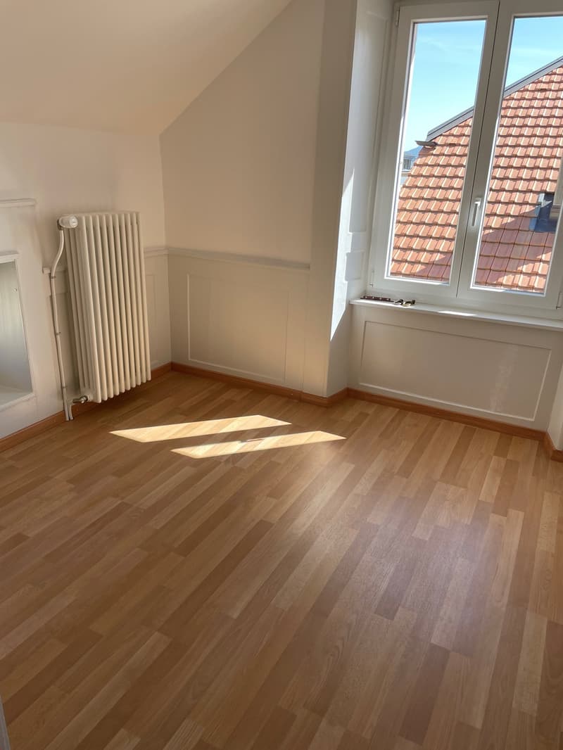 large empty room with white walls, wooden floor, large window with sunrays coming in, heater on the wall