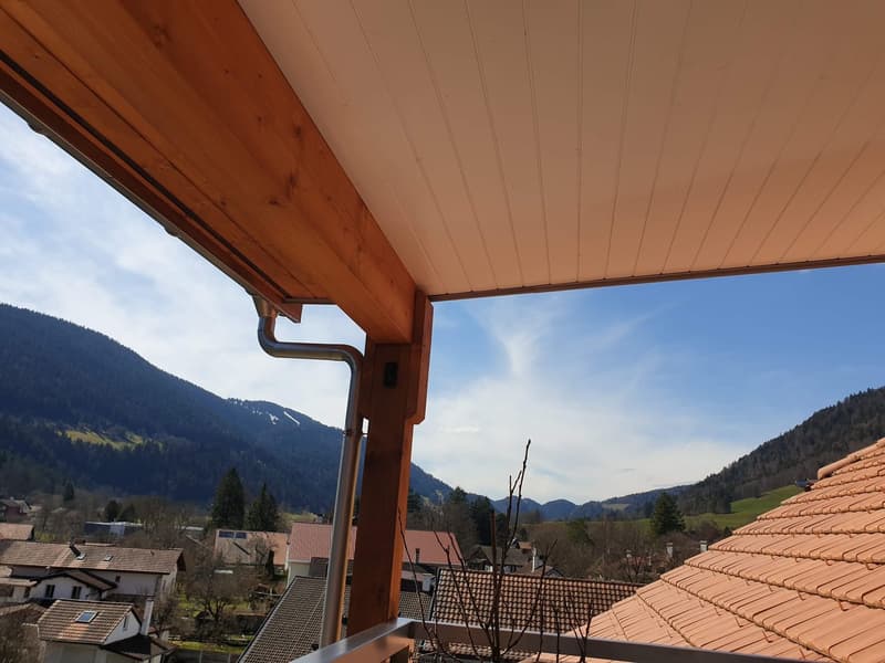 View from a wooden balcony with a metal railing overlooking houses, hills, and mountains