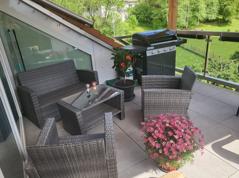 a glass walled balcony, with a sitting area, a table, and a barbecue. Surrounded by trees and a green valley.