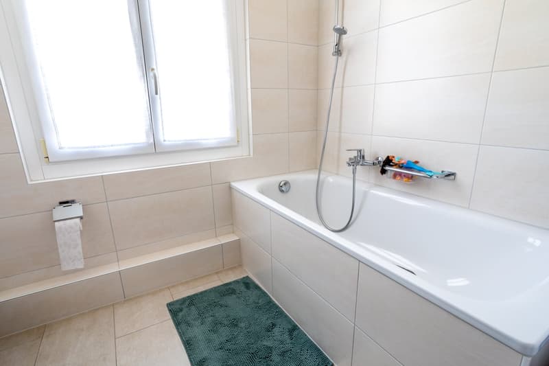 Bathroom with bathtub, shower, window, and tiled walls and floor.