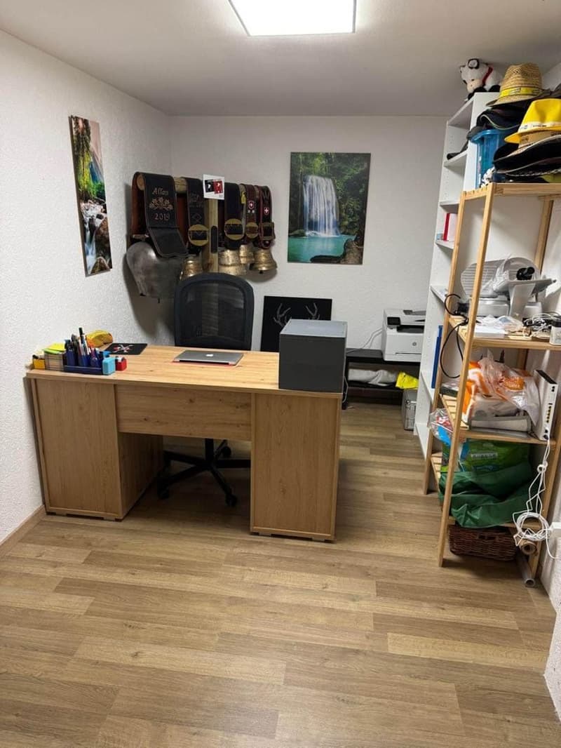 wooden desk, black chair, wooden floor, shelf with books and items, paintings on wall, and a laptop on desk