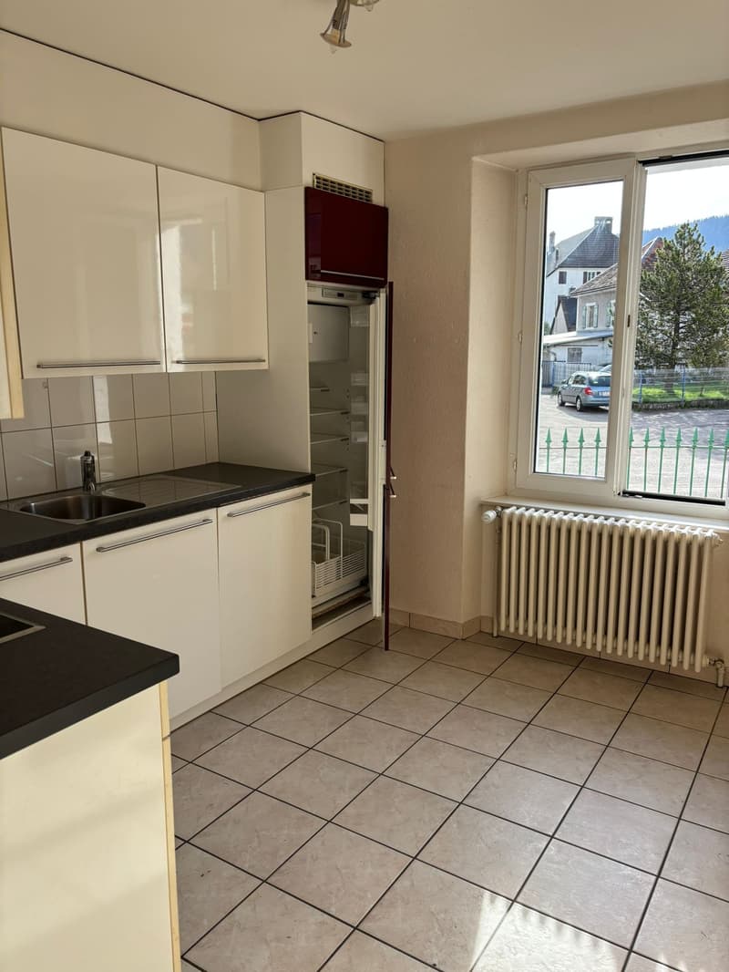Modern kitchen with white cabinets, black countertops, stainless steel sink, and a refrigerator. Tiled floor and a radiator under the window.