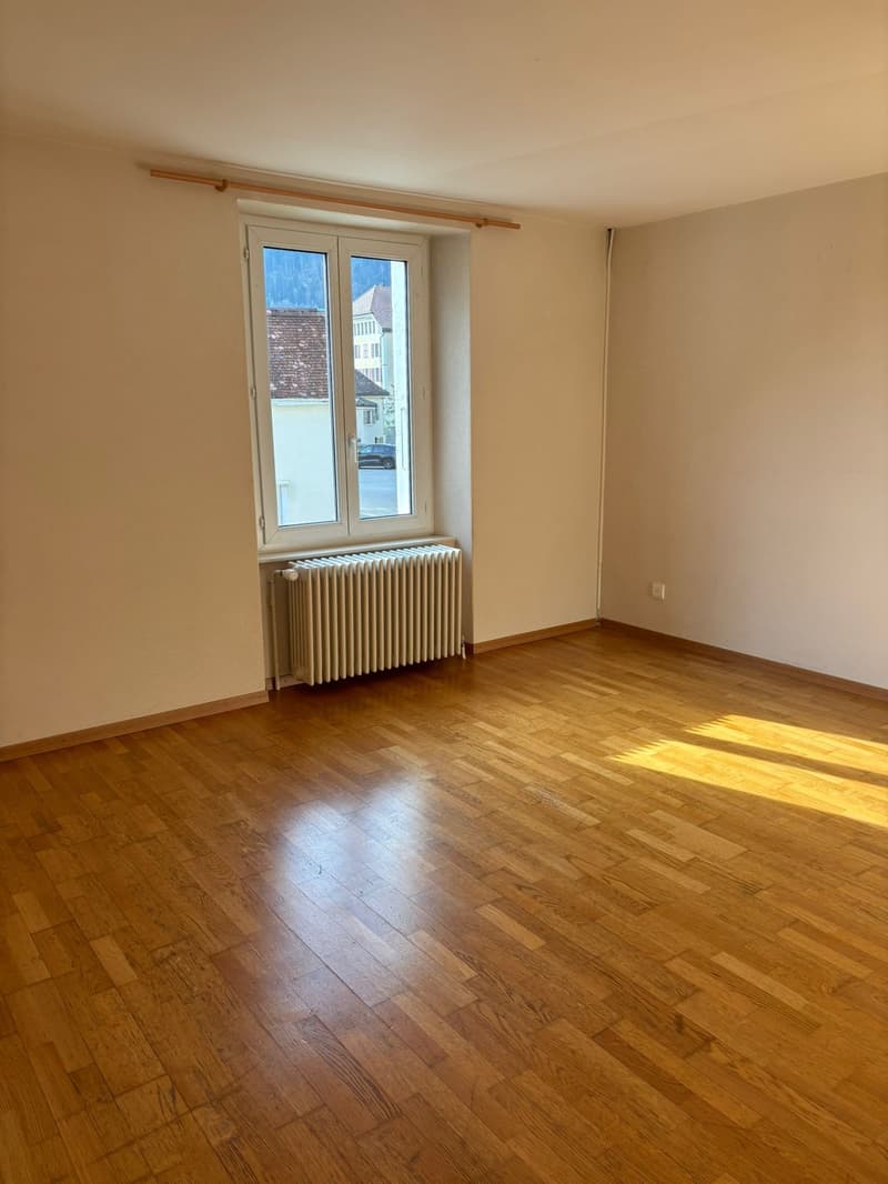 An empty room with wooden floors, white walls, a window with a view of the outside, and a white radiator.