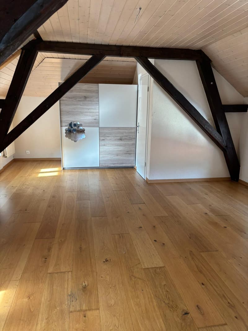 An empty room with wooden floors and exposed wooden beams on the ceiling. The room has a white wall and a white door. There is a white cabinet with a mirror on the wall. The room is illuminated by sunlight coming from the windows.
