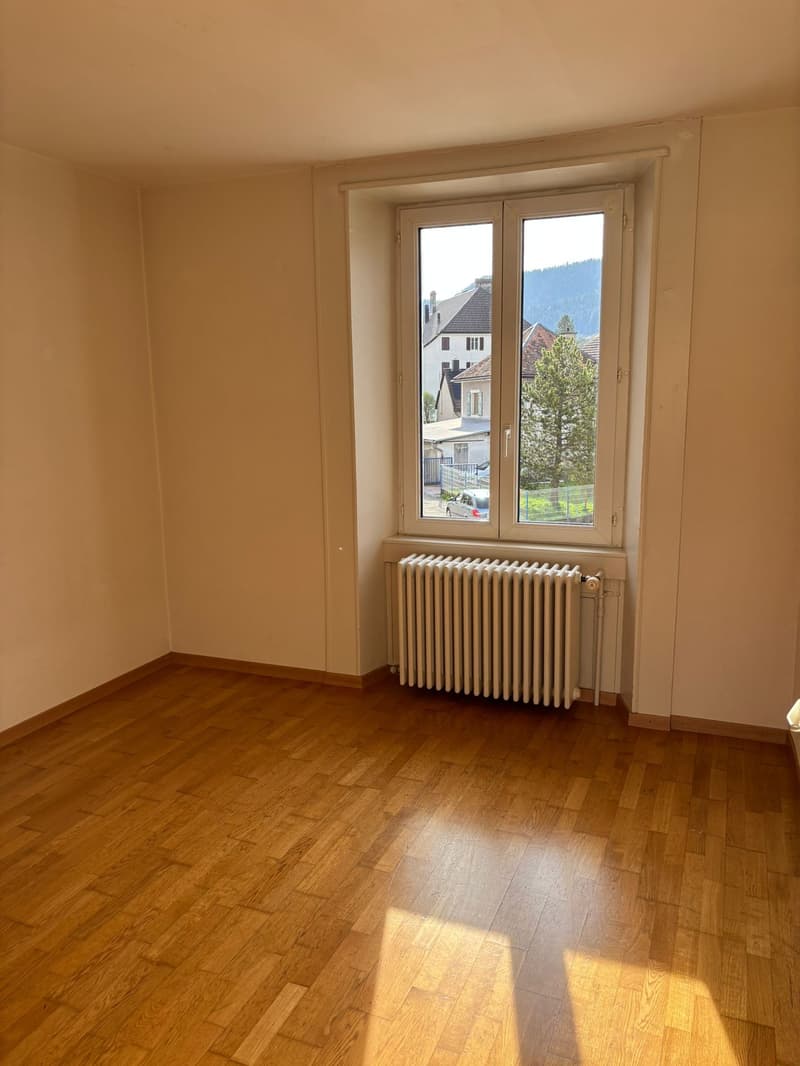 Empty room with wooden floor, white walls, and a large window with a view of a house and trees outside. A white radiator is installed below the window.