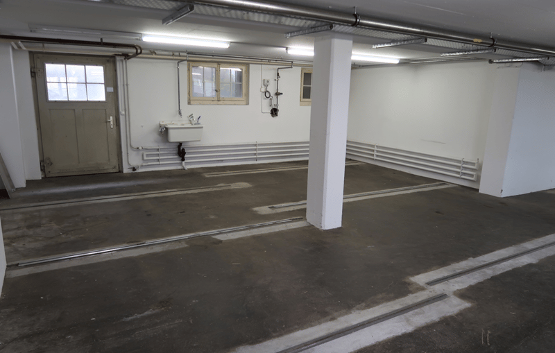 Empty concrete floor, concrete walls, white sink, overhead lighting, pipes on ceiling