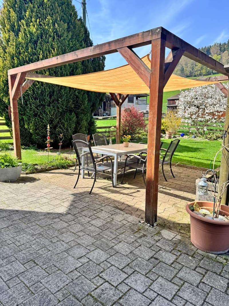 Outdoor dining area with a wooden pergola, table, chairs, and a potted plant.
