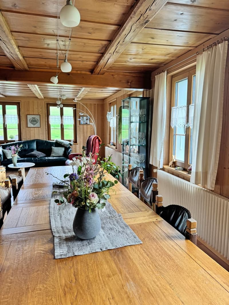 large wooden dining table, flowers in vase, wooden chairs, black leather couch, wooden ceiling, windows