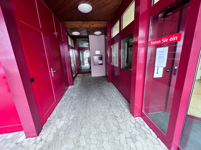 The image shows a hallway with bright pink walls and a wooden ceiling. The floor is covered in gray hexagonal tiles. There are glass doors leading to the outside, and a sign on the wall that says 'Treten Sie ein' which means 'Enter' in German.