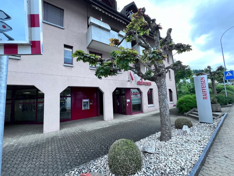 A multi-story commercial building with a red and white exterior, featuring a Raiffeisen bank branch on the ground floor. The building has large windows and a tree growing in front of it, surrounded by gravel landscaping.