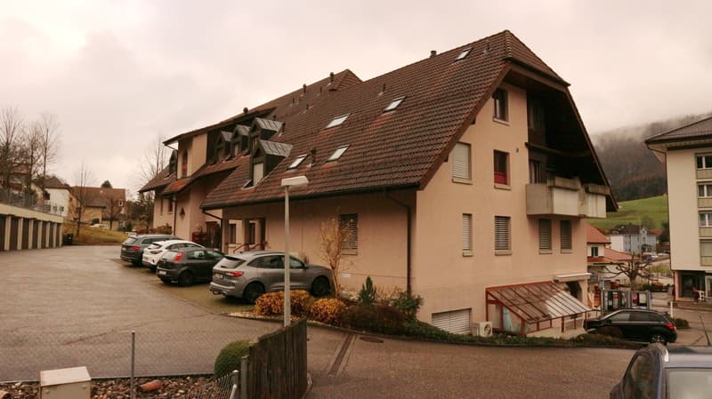 Multi-story apartment building with red tile roof, tan exterior walls, and several windows. There is a parking area in front of the building with several cars parked.
