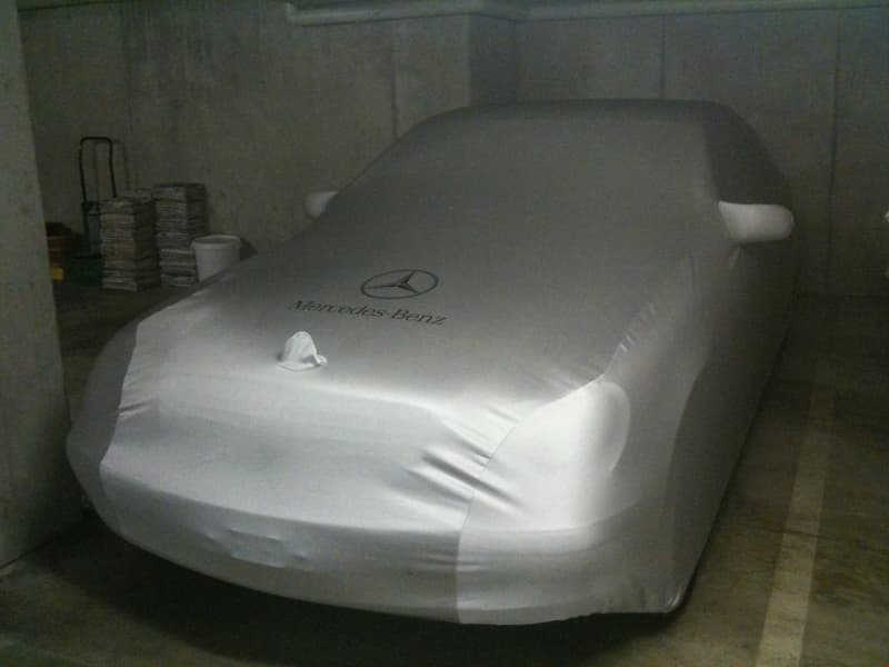 A covered Mercedes-Benz car parked in a garage or storage area. The car is covered with a protective cover, likely to keep it clean and protected when not in use.