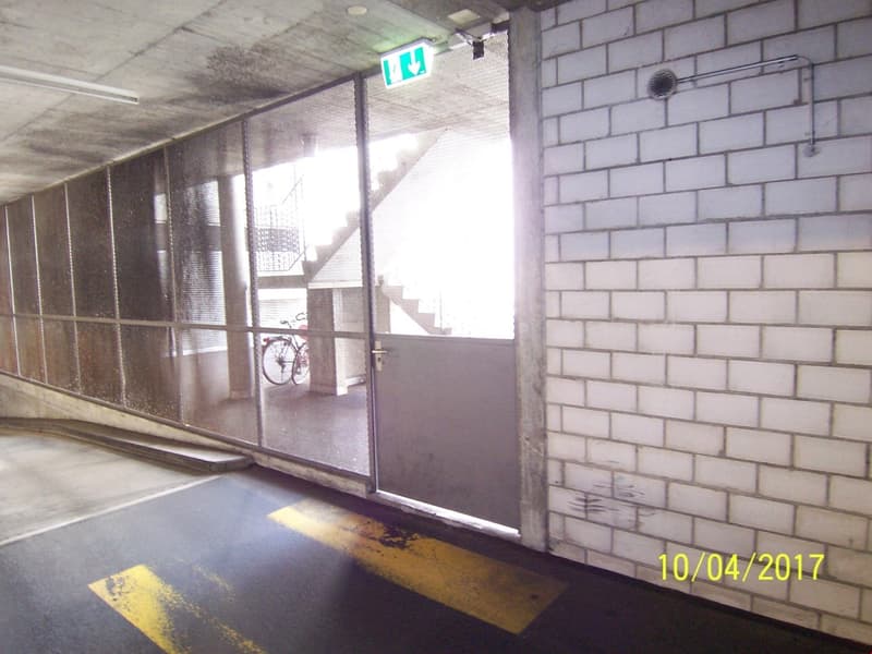 The image shows a hallway or corridor inside a building. The walls are made of white bricks, and there are large windows along one side. There is a green exit sign visible on the ceiling, and the floor appears to be tiled or made of some other hard materi