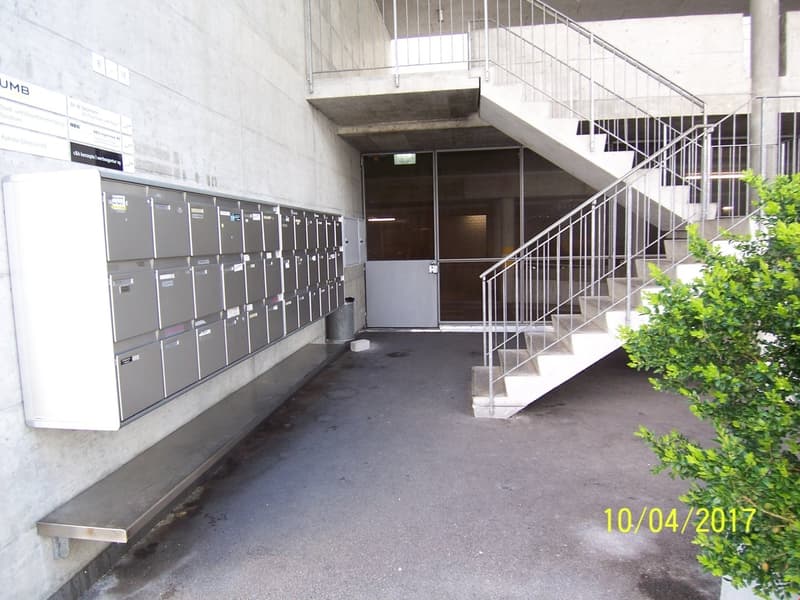 The image shows the entrance to a building, with a set of metal lockers or mailboxes on the left side and a staircase leading up to the entrance on the right. The entrance appears to be made of concrete and has a metal railing. There are some plants visib