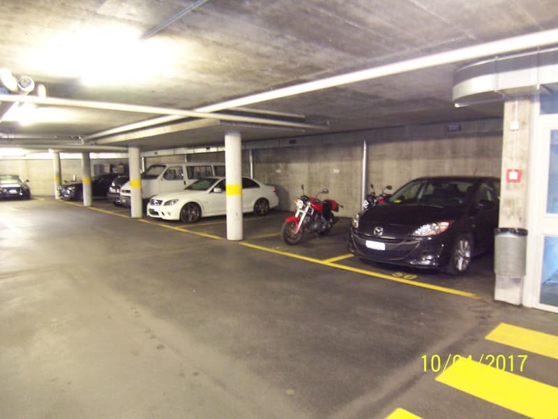 Underground parking garage with several cars and a motorcycle parked in designated spots. The garage has a concrete ceiling and floor, with yellow safety lines on the ground.