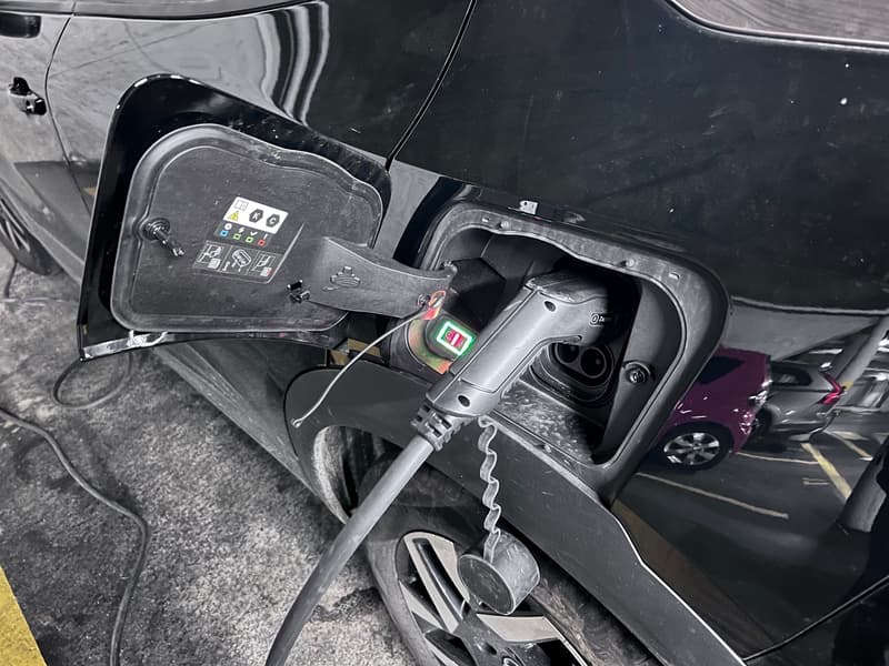 The image shows the charging port of an electric vehicle. The charging port is located on the side of the vehicle and has various indicators and controls. The vehicle appears to be parked in a parking lot or garage.
