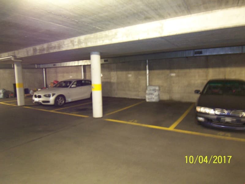 Underground parking garage with concrete walls and floor, yellow parking lines, and several parked cars including a white BMW and a dark-colored SUV.