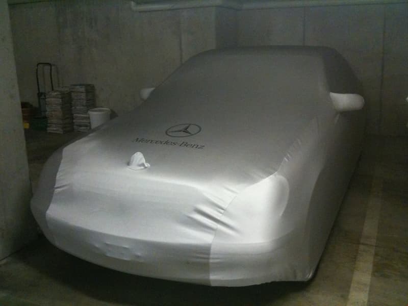 A covered Mercedes-Benz car parked in a garage or storage area. The car is covered with a white protective cover, and there are some stacked items visible in the background.