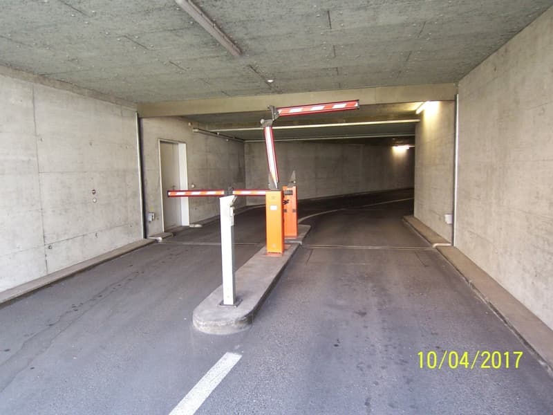 This image shows the interior of a parking garage. The space has concrete walls and floor, with a raised barrier and parking space markings visible. There is a yellow parking barrier and overhead lighting fixtures in the space.
