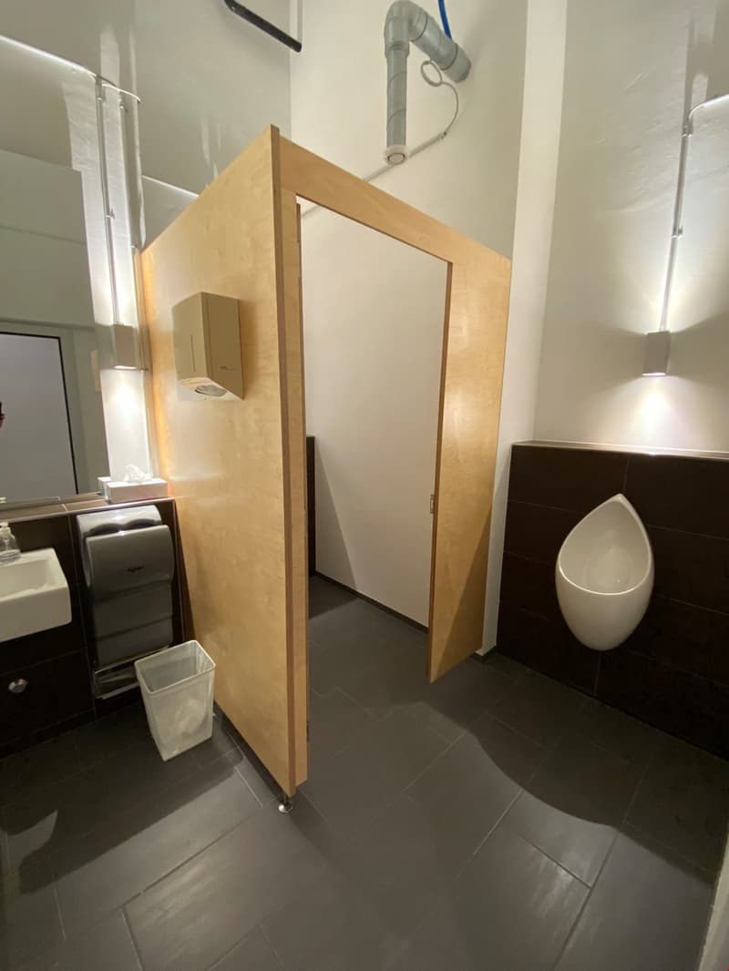The image shows a bathroom with a wooden cabinet, a urinal, and a washing machine. The floor is tiled in a dark color, and the walls are light-colored. There is also a ventilation system visible in the ceiling.