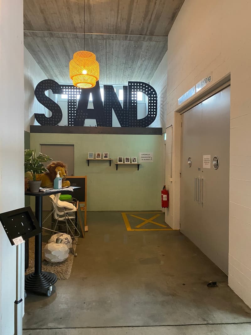 The image shows an office space with a large 'SAND' sign on the wall, a desk with a computer and other office supplies, and a yellow lamp hanging from the ceiling. The walls are made of concrete and the floor is a concrete surface. There is also a fire ex