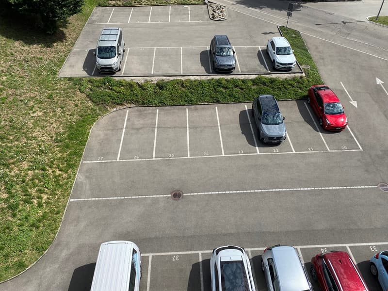 Aerial view of a parking lot with numbered parking spaces, several cars parked, and a grassy area with shrubs on the side.