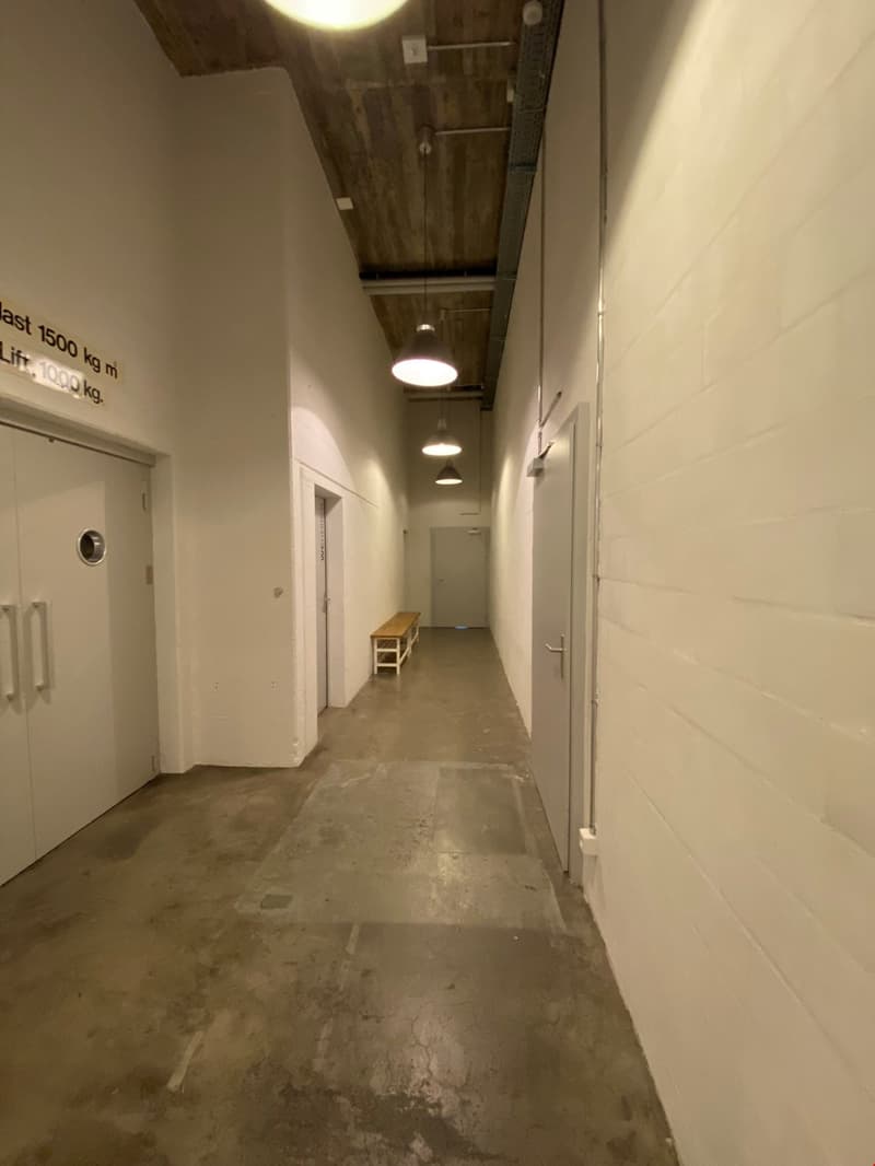 Narrow hallway with concrete floor, white walls, and recessed lighting. Wooden ceiling beams are visible. There is a bench or small table against the wall and several doors leading to other rooms.