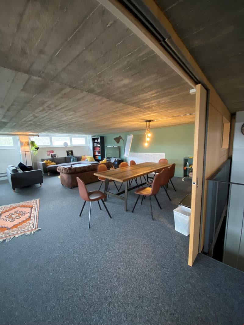 The image shows an open-plan living and dining area with a concrete ceiling, hardwood dining table and chairs, a sofa, and shelving units. The space has large windows that provide natural light and access to a balcony or terrace.