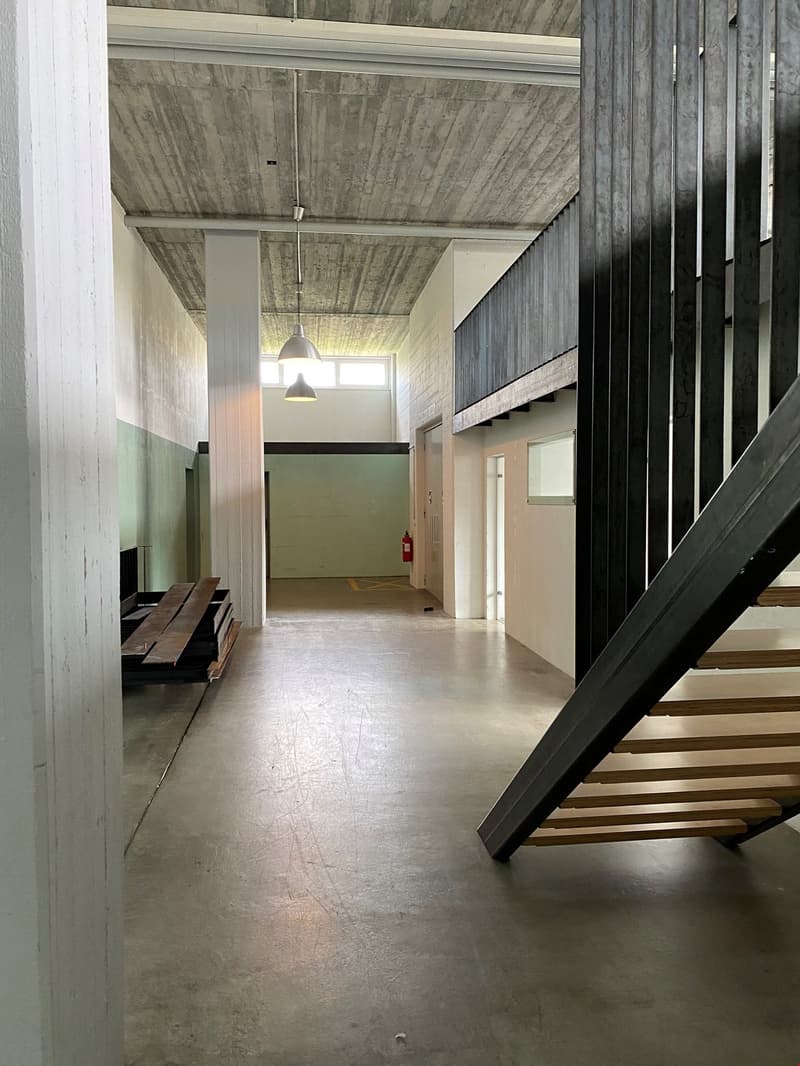 The image shows the interior of a building with a concrete ceiling and walls, and a wooden staircase. The space appears to be a hallway or entryway, with a door visible in the background. The floor is made of a concrete-like material, and there is a light