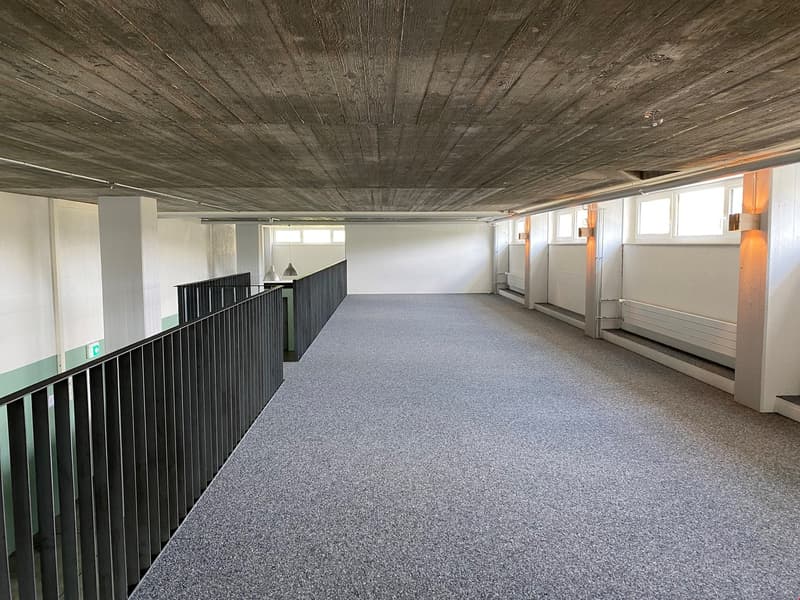 The image shows a large open space with a high ceiling and a wooden-paneled ceiling. The floor is covered in gray carpeting, and there are black metal railings along the upper level. The walls are white, and there are several windows providing natural lig