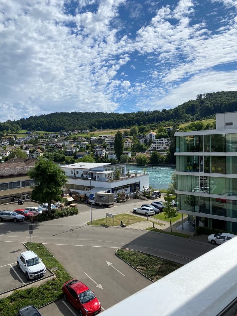 The image shows a scenic view of a town or city nestled in a valley, with a river or stream running through it. The buildings are a mix of residential and commercial structures, with some modern glass-fronted buildings visible. The surrounding hills are c