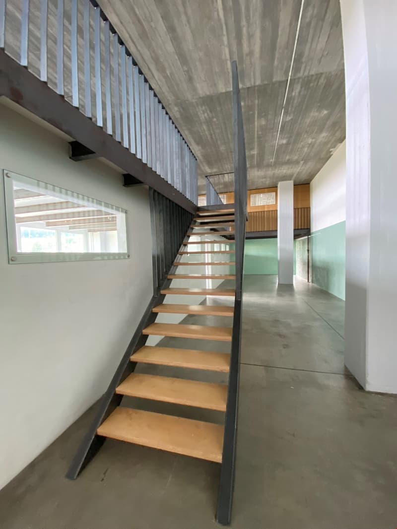 The image shows a modern interior space with a concrete ceiling and walls, and a wooden staircase leading to an upper level. The staircase has a metal frame and wooden steps. The floor appears to be a concrete surface. The space has a minimalist and indus