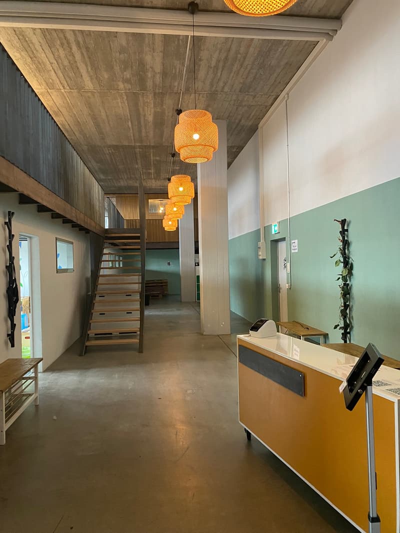The image shows the interior of a building with a modern, minimalist design. The space features a wooden ceiling with exposed beams and pendant lighting fixtures with a warm, amber glow. The walls are a mix of neutral tones and a light mint green color. T