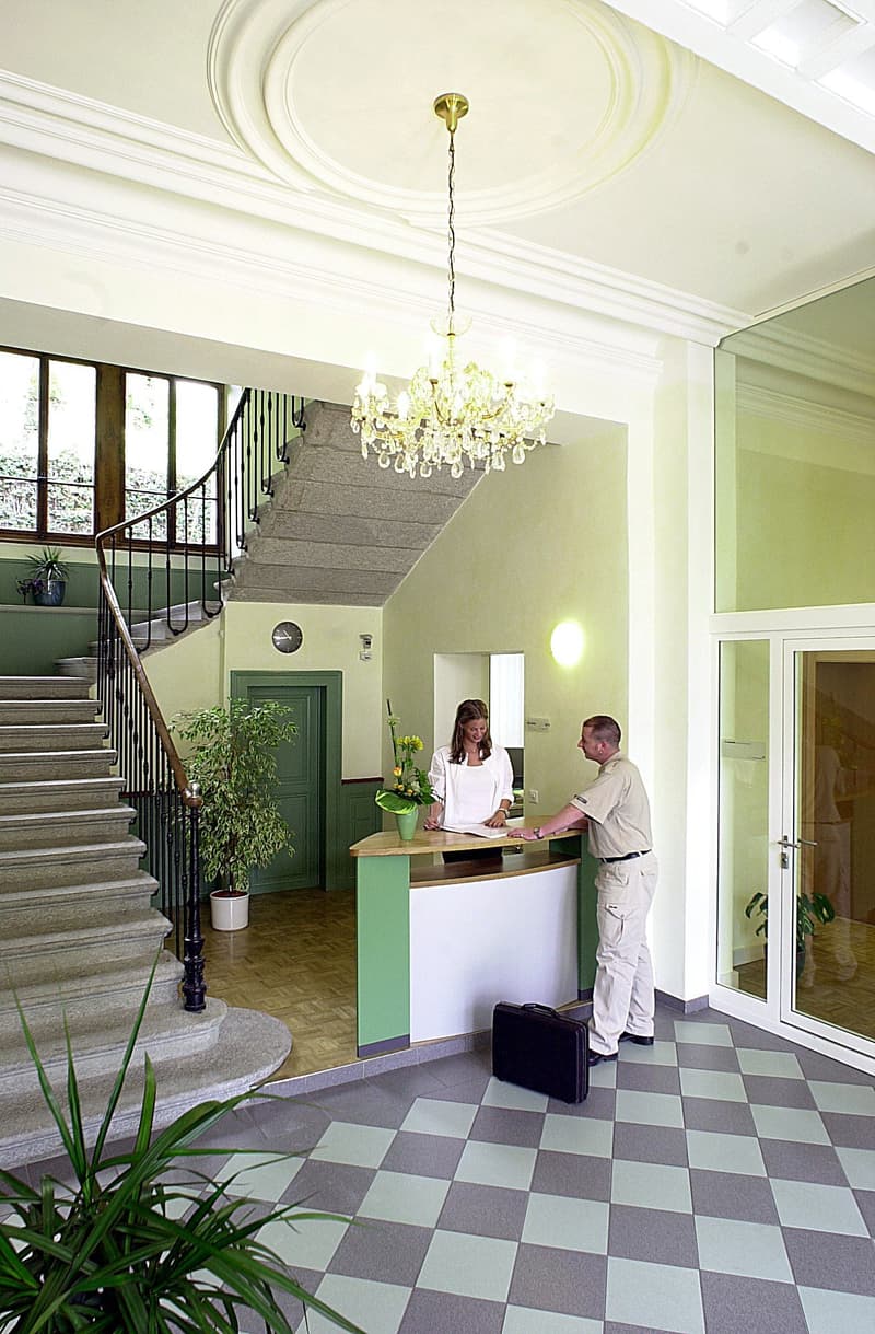 Spacious lobby with a grand staircase, a chandelier, and a reception desk. The floor is tiled in a checkered pattern, and there are potted plants decorating the space. Two people are visible at the reception desk.