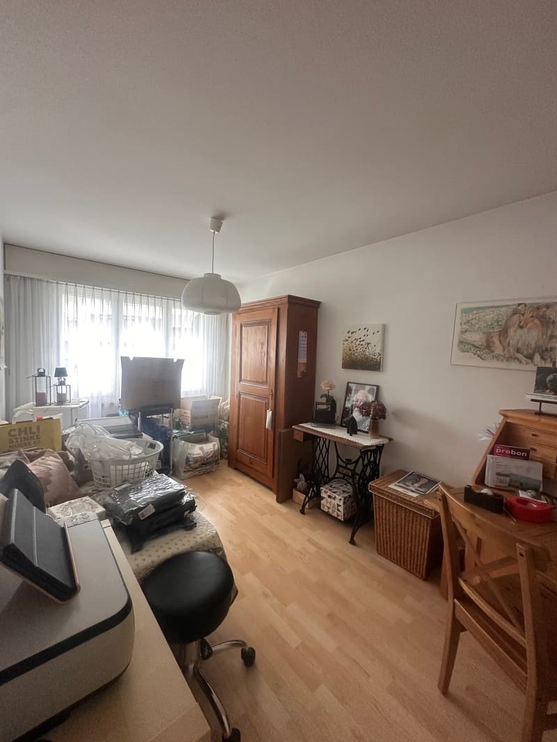 living room with wooden floor, white walls, wooden cabinet, desk, chair, table, lamp, window, curtains, picture frame, flower vase, printer, stool, storage boxes, baskets, boxes, and other items