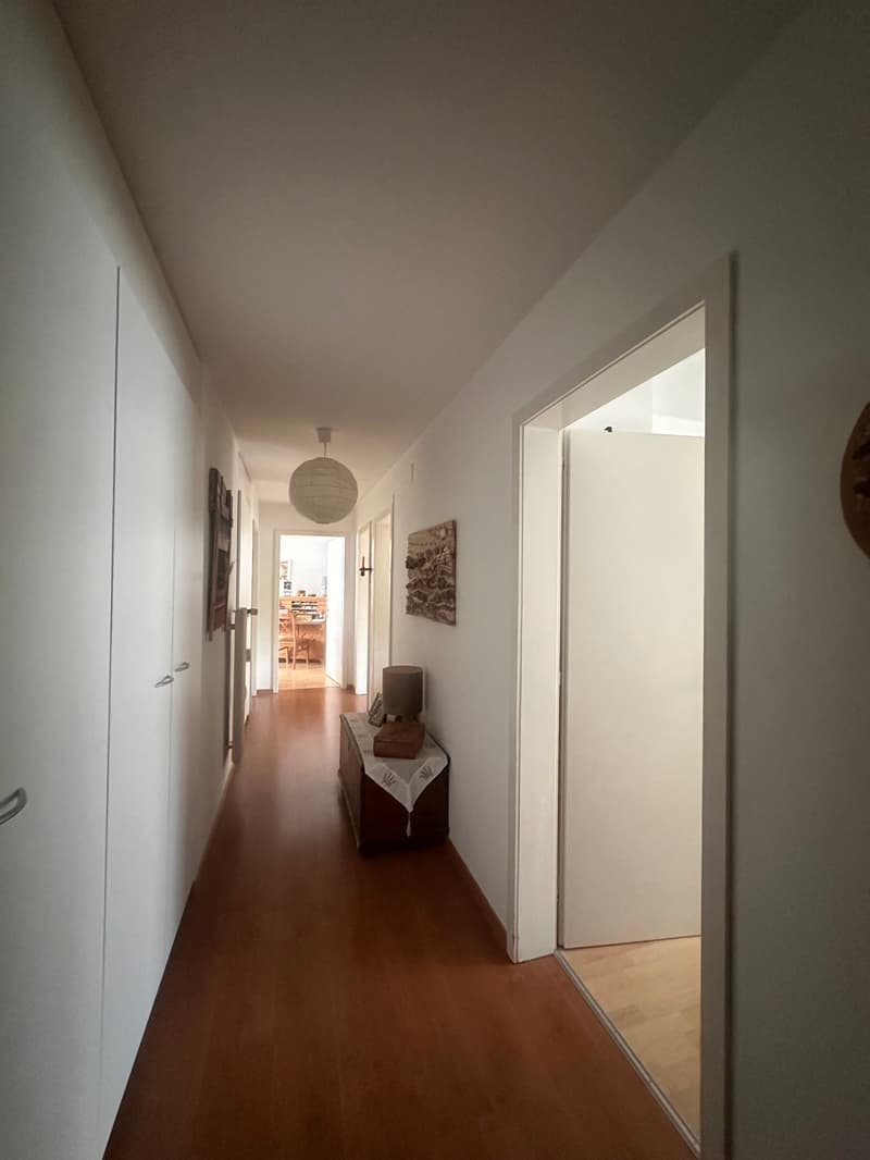 long hallway, wooden floor, white walls, white doors, lamp, table with lamp, picture frame, artwork on wall