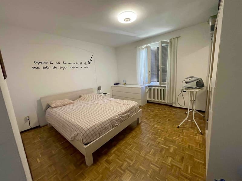 bedroom with white walls, wooden parquet floor, bed with checkered bedspread, night lamp, small table, whiteboard with text, open window with blinds, radiator, white ironing board, white cabinet