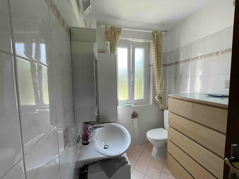 White tiled walls, white tiled floor, sink with faucet, toilet, window with curtains, wooden drawer unit, mounted soap dispenser, air vent on ceiling