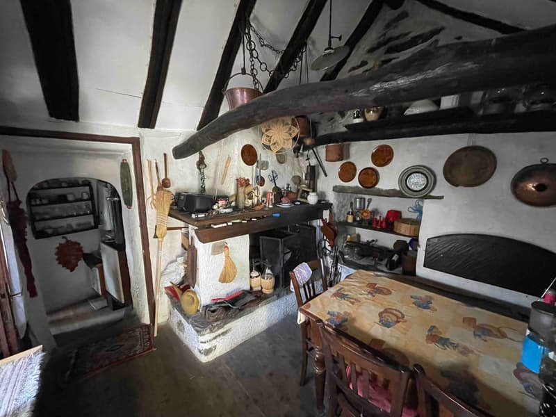 stone walls, wooden ceiling, large fireplace, dining table, wooden chairs, kitchen shelves, vintage objects, brooms, baskets, pots, pans