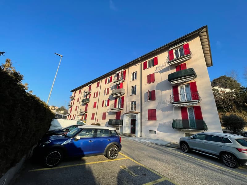 White multi-story building with red shutters, 3 parked cars, bushes, street light