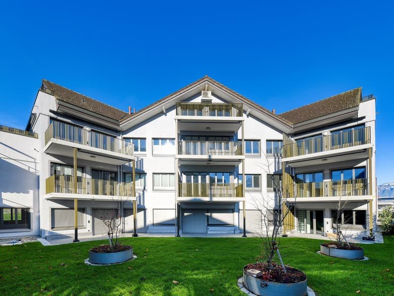 White multistory building, brown roof, large balconies, large windows, green front yard, metal planters